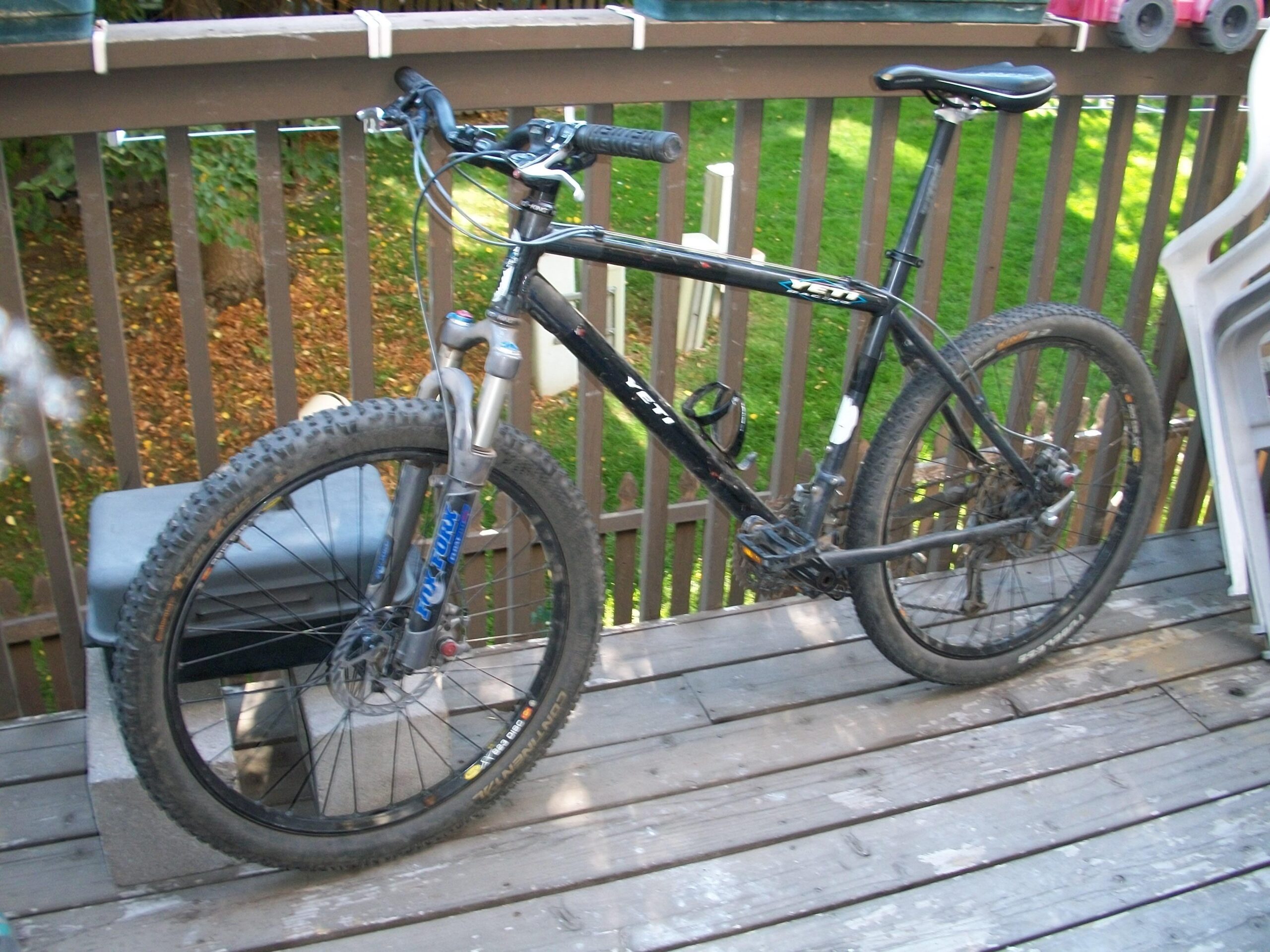 Kona Caldera: A black mountain bike with dirt-covered tires is resting on a wooden deck. The bike features a front suspension fork and has a few visible markings of wear. Behind it, there is a green lawn and some trees, creating a natural background. Nearby, there is a gray storage box made of cinder blocks.