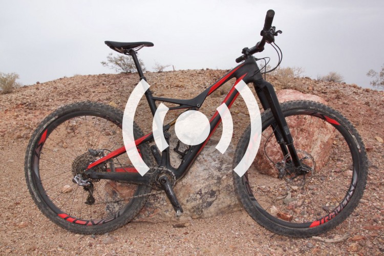 A black and red mountain bike leaning against a rocky surface, set against an outdoor landscape with a gravel and dirt background. The bike features thick tires suitable for rugged terrain and has a sleek design with visible gears and components.