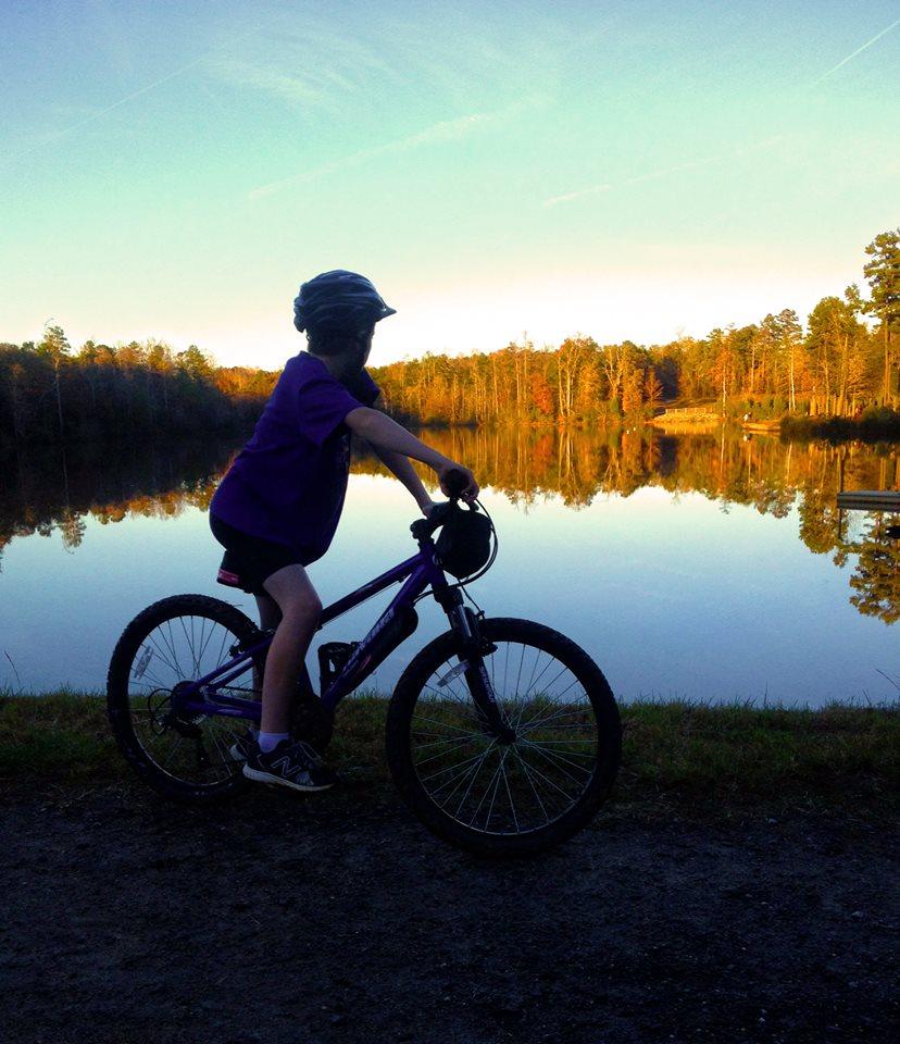 A child in a purple shirt and helmet is riding a bicycle along a scenic path near a calm lake. The water reflects the colorful trees and the clear sky, creating a peaceful autumn atmosphere. Park-rd. Short Trails. mountain bike trail.
