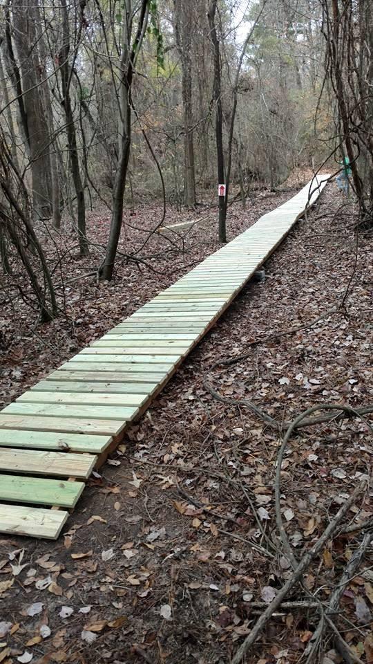 A wooden boardwalk extending through a wooded area, surrounded by bare trees and scattered leaves on the ground. The path is straight and narrow, leading further into the forest. Bringle Lake Mountain Bike Trail System mountain bike trail.