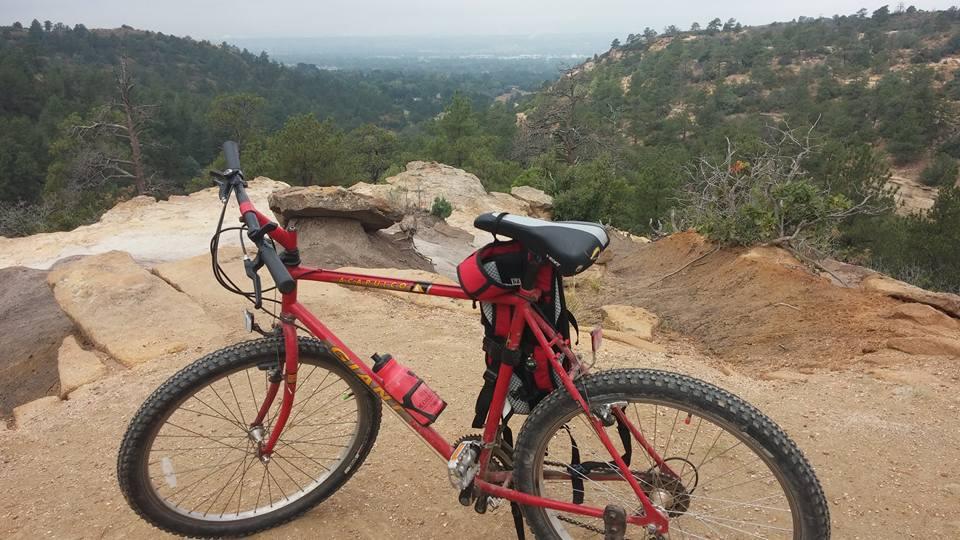 A red mountain bike parked on rocky terrain, with a scenic view of green hills and valleys in the background under an overcast sky. Palmer Park mountain bike trail.