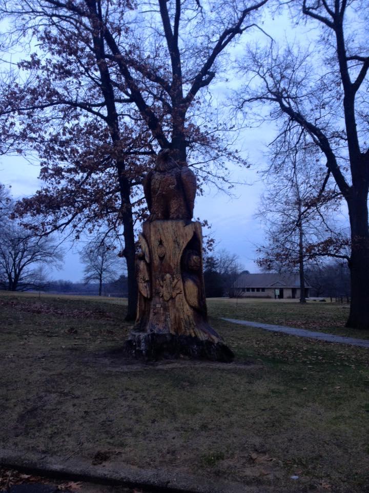 A large, intricately carved wooden sculpture of an owl perched atop a tree stump, surrounded by bare trees and a grassy area. The sky appears overcast, giving the scene a slightly gloomy atmosphere. In the background, a building is partially visible, adding context to the outdoor setting. Brighton Rec Area mountain bike trail.