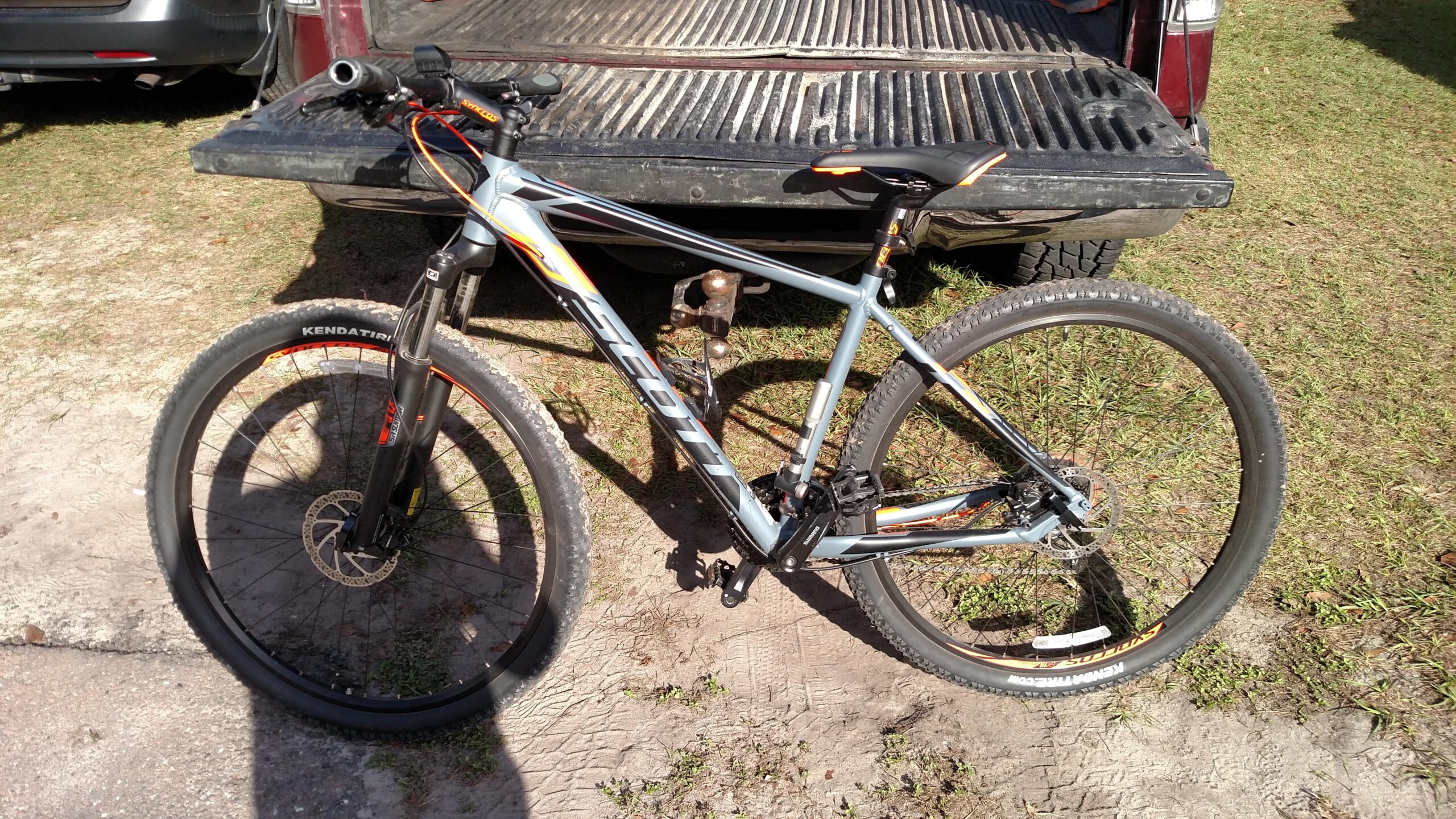 Scott Aspect 950: A silver and orange mountain bike is parked on a grassy area next to the tailgate of a truck. The bike features thick tires and a front suspension fork. The background shows part of the truck's bed and another vehicle parked nearby.
