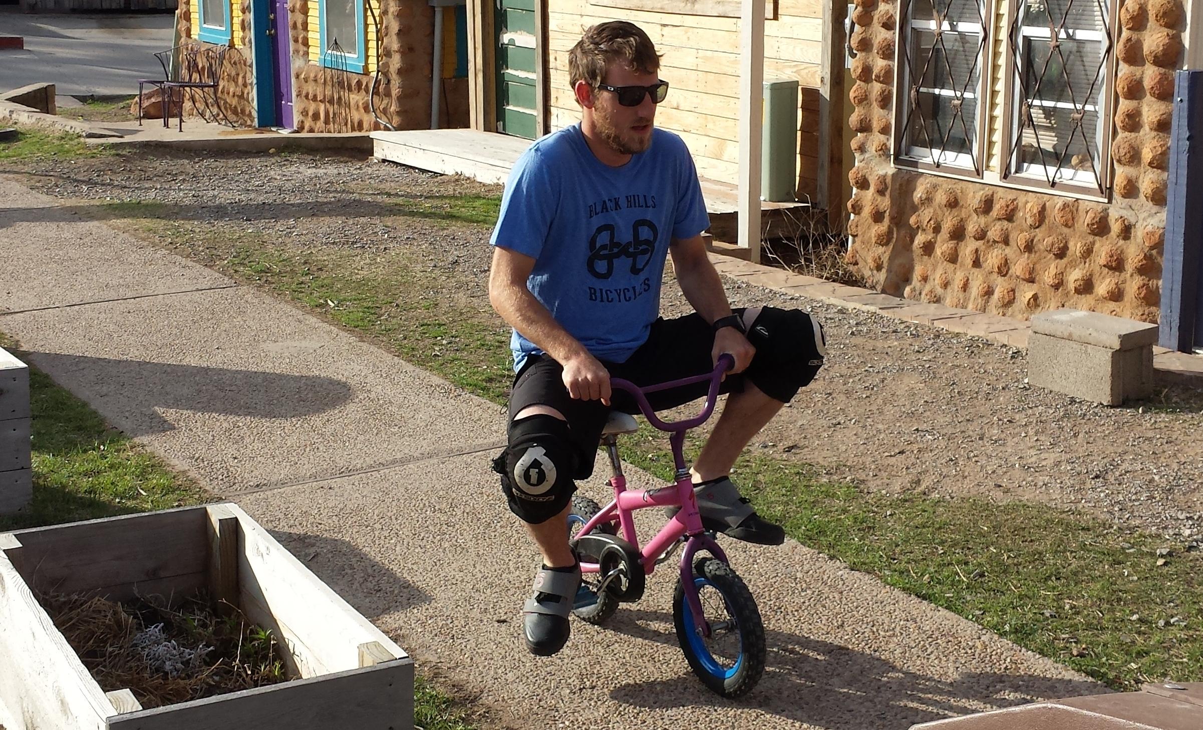 A man wearing sunglasses and a blue shirt is riding a small pink bicycle on a pathway. He has knee pads on and is positioned in a casual riding stance. In the background, there are colorful buildings and a patch of grass with wooden planters. Lake Lawtonka Trails mountain bike trail.