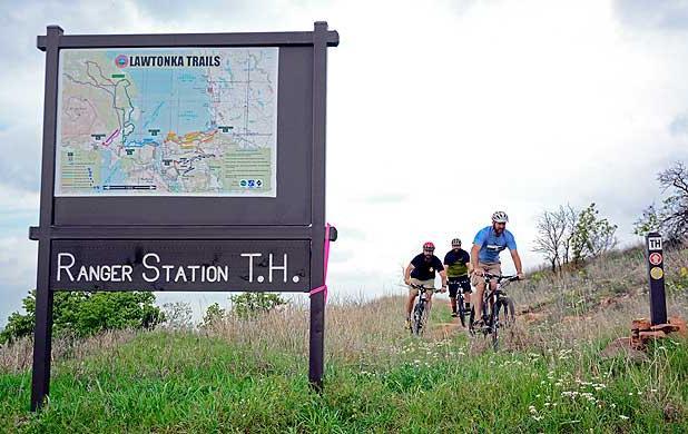 Three mountain bikers are riding along a trail near a sign that reads "Ranger Station T.H." The sign features a map of the Lawtonka Trails. The landscape is grassy with a few trees, under a partly cloudy sky. Lake Lawtonka Trails mountain bike trail.