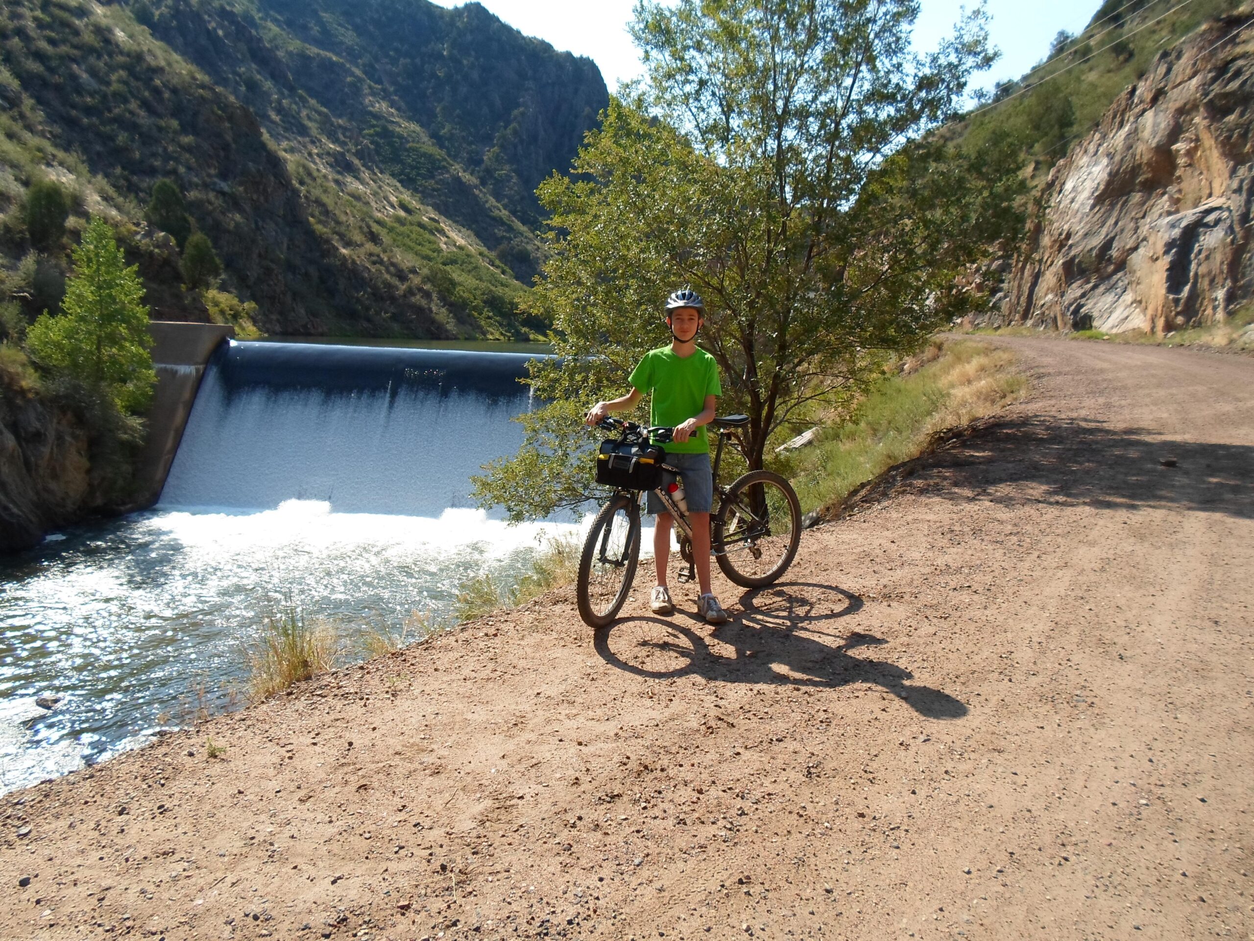 Trek 820: A young person in a green t-shirt and helmet stands next to a bicycle on a dirt path beside a river. In the background, a dam releases water into the river, with rocky hills and greenery surrounding the scene. The sun is shining brightly, creating a lively outdoor atmosphere.