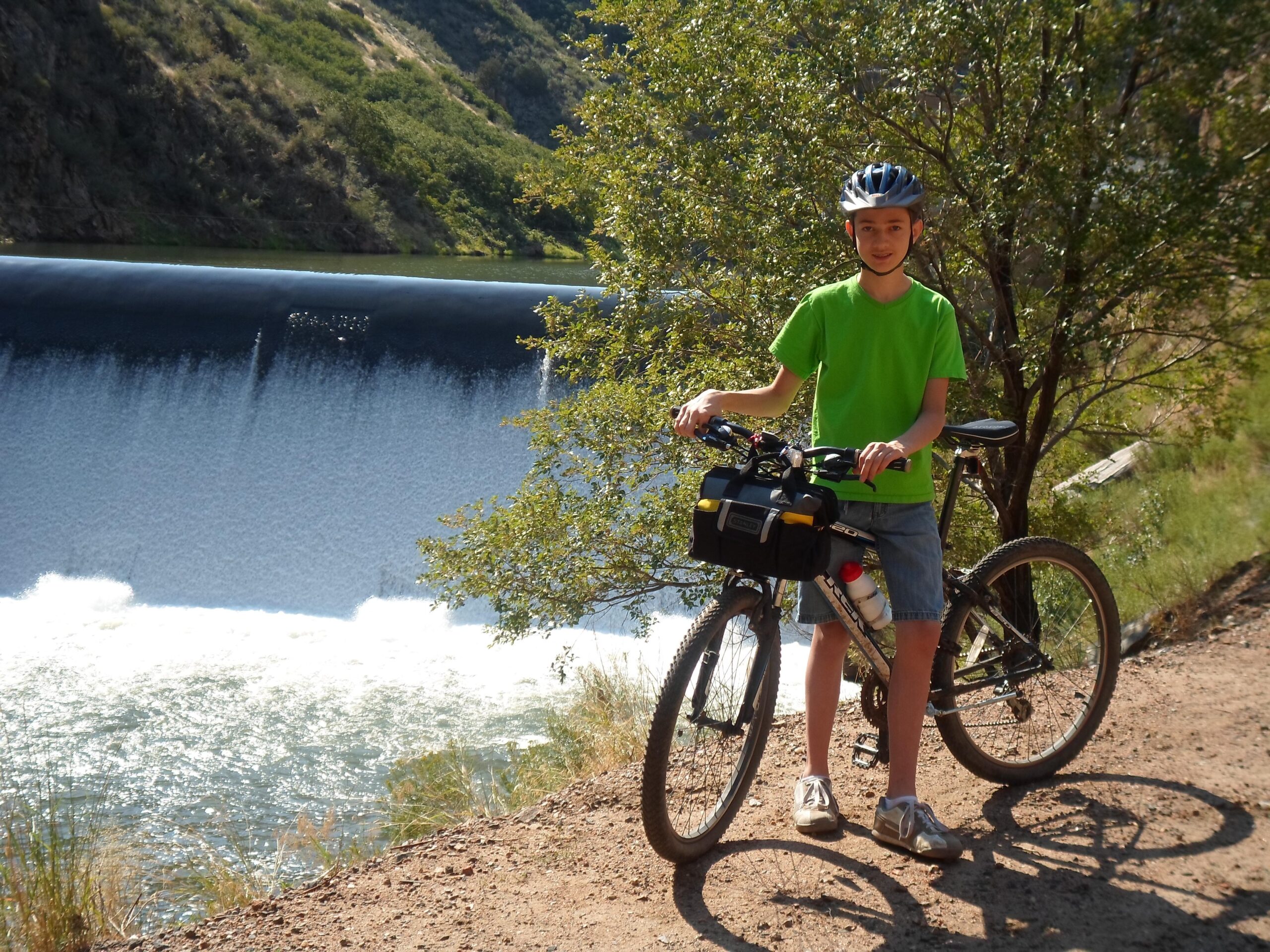 Trek 820: A young boy wearing a green t-shirt and helmet stands beside his mountain bike on a dirt path, with a scenic view of a body of water and a dam in the background. Trees and hills surround the area, suggesting a sunny outdoor environment.