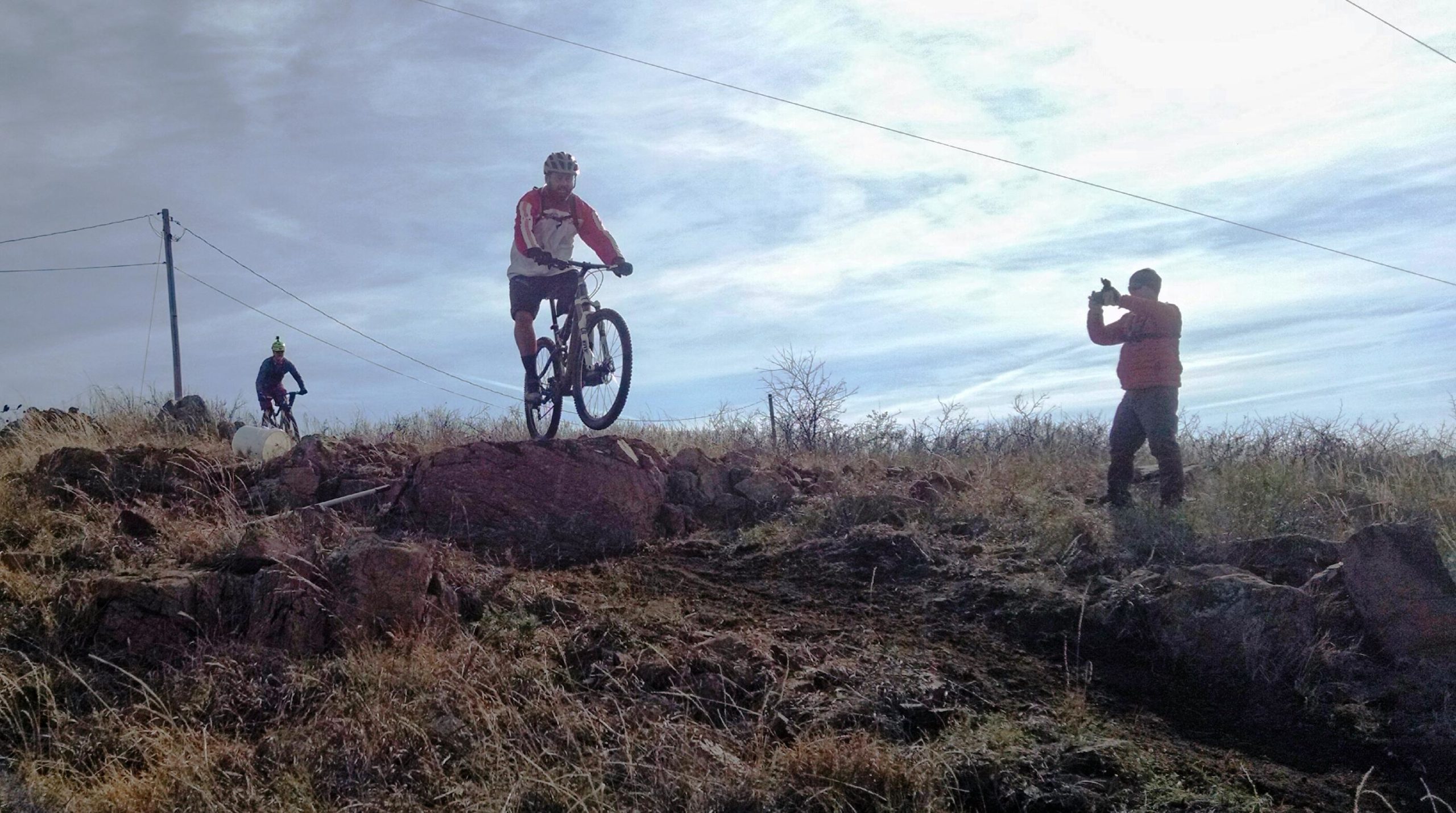 A mountain biker performs a jump over a rocky outcrop while another person stands on the hillside, photographing the action. The scene is set in a natural outdoor environment with sparse grass and scattered rocks under a cloudy sky. Lake Lawtonka Trails mountain bike trail.