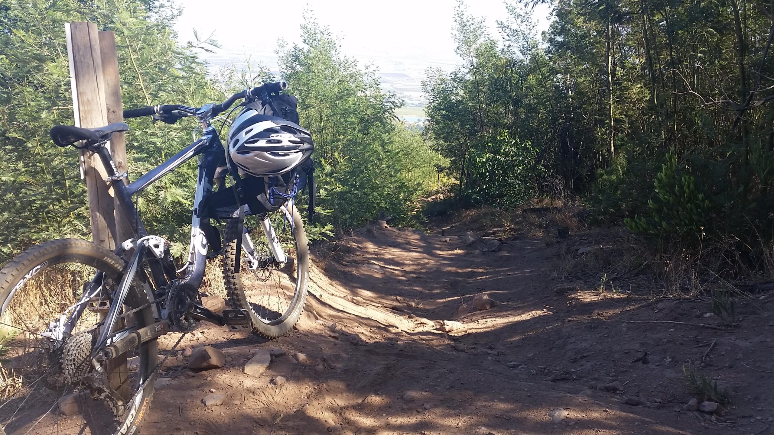 Giant Trance: A mountain bike is parked next to a wooden post on a dirt trail surrounded by trees. The trail slopes downward, indicating a rugged terrain ideal for biking. A helmet is attached to the bike, and the scene captures a lush, natural environment.