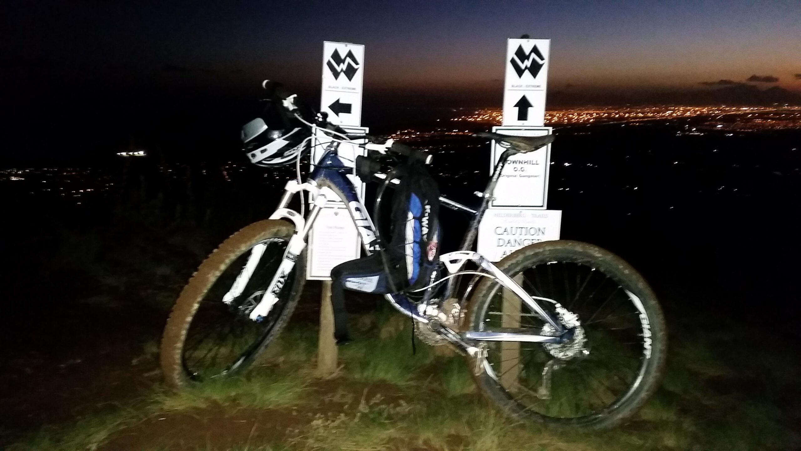Giant Trance: A mountain bike with a backpack rests against trail signs at dusk, overlooking a city illuminated by lights in the background. The signs indicate different trail options, with one pointing downhill and warning of potential dangers. The scene captures the adventurous spirit of mountain biking as day transitions to night.