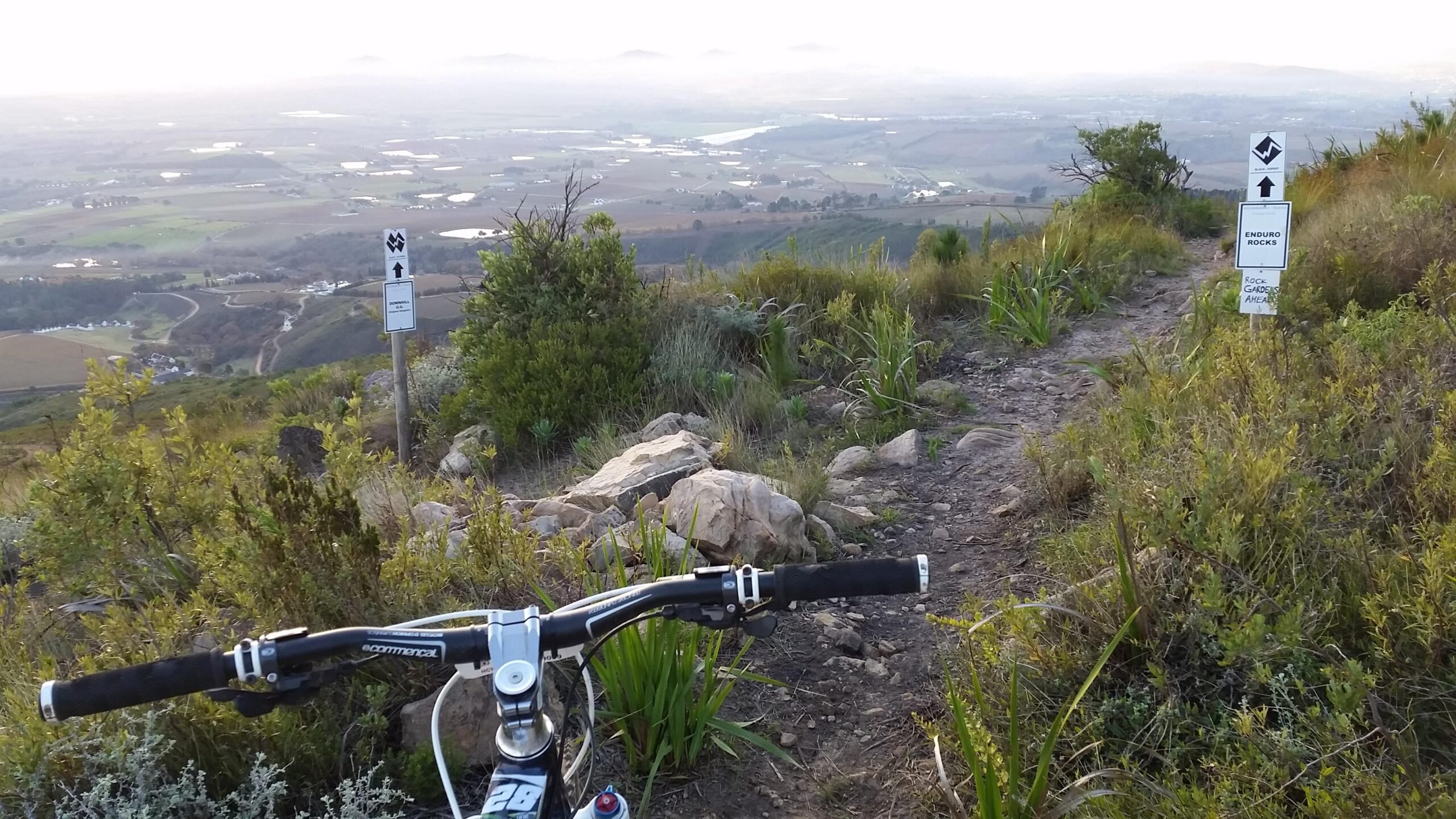 Giant Trance: A mountain bike is positioned on a rocky trail overlooking a valley filled with farmland, rivers, and distant mountains in the background. Trail markers are visible alongside the path, indicating upcoming terrain challenges. The scene captures the natural beauty of the landscape at dusk.