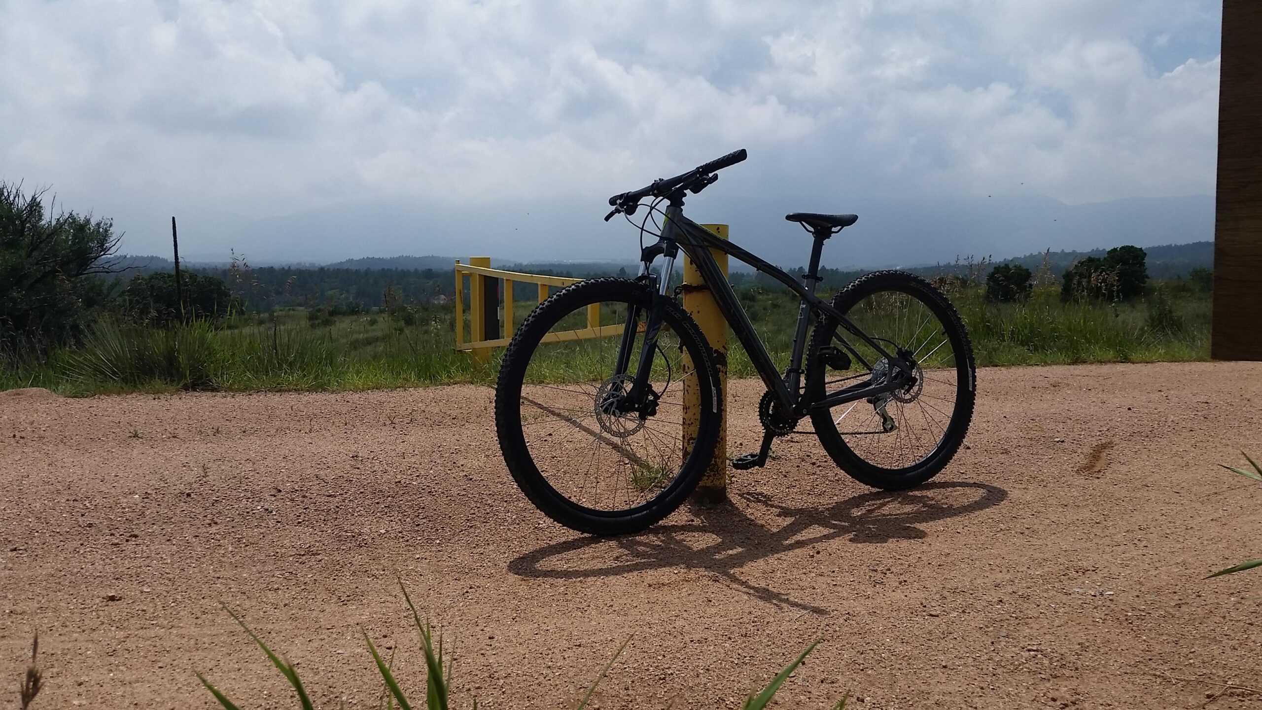Specialized Rockhopper 29: A black mountain bike parked next to a yellow gate on a gravel path, with a green landscape and cloudy sky in the background.