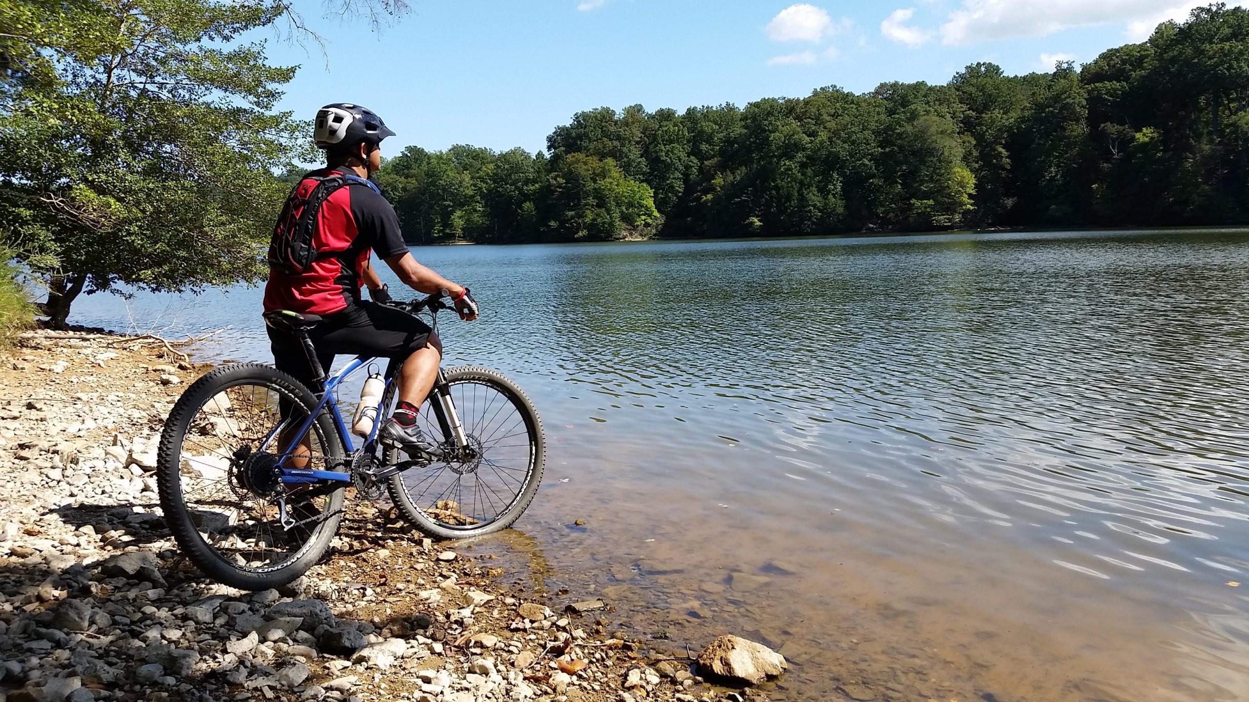 A person wearing a helmet and a red and black shirt is sitting on a mountain bike beside a calm lake, looking out over the water. The scene features rocky terrain, trees in the background, and a clear blue sky with a few clouds. Fountainhead Regional Park mountain bike trail.