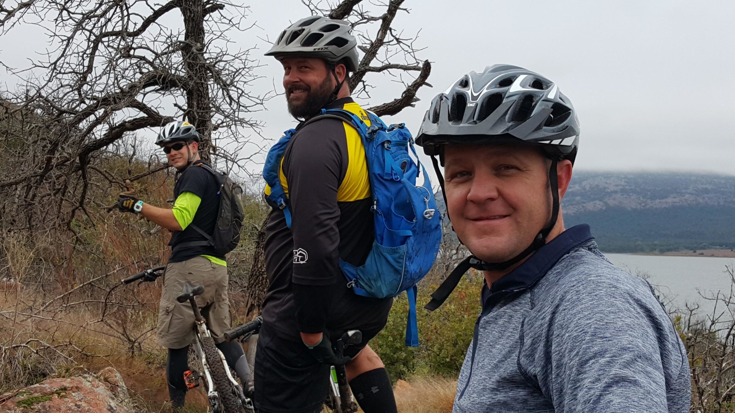 Three mountain bikers pose for a photo on a trail, surrounded by sparse trees and a scenic landscape in the background. They are wearing helmets and cycling gear, with two of them smiling at the camera. The setting appears to be outdoors, possibly near a body of water, under an overcast sky. Lake Lawtonka Trails mountain bike trail.