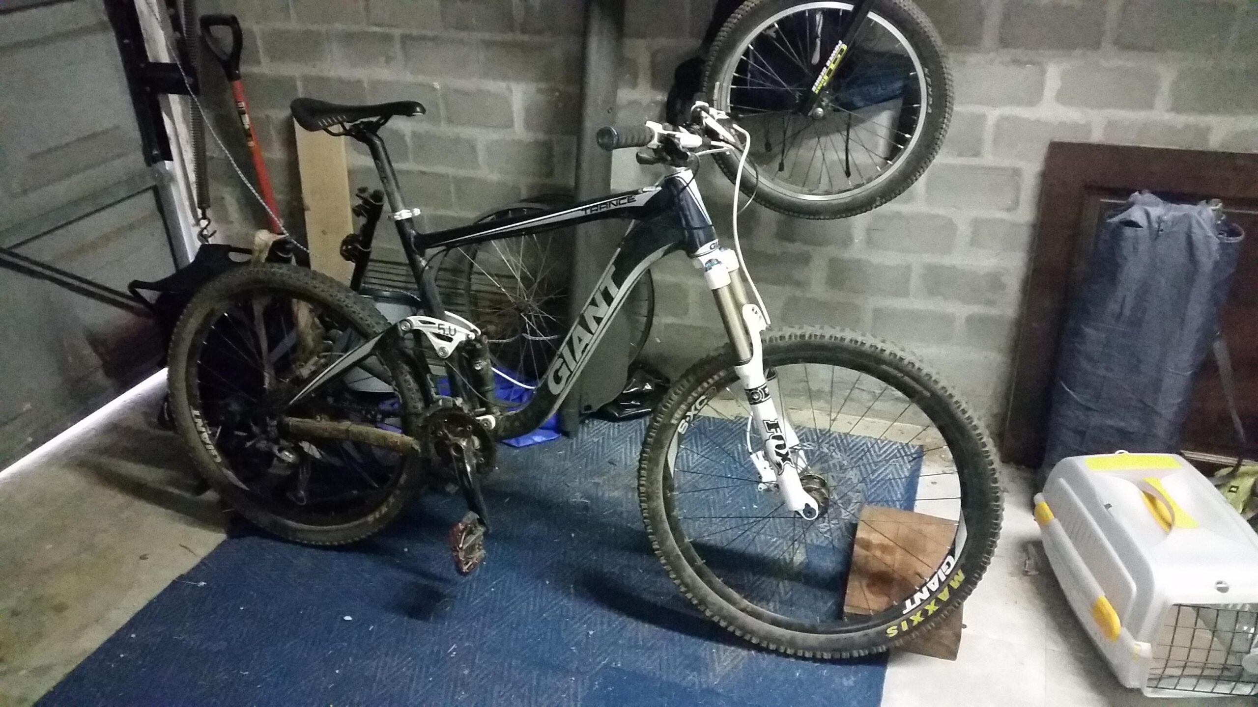 Giant Trance: A mountain bike leaning against a wall in a garage, with a black frame and visible branding. The front wheel is raised, and there are some tools and a pet carrier in the background. The floor features a rubber mat, and the garage has a concrete wall.