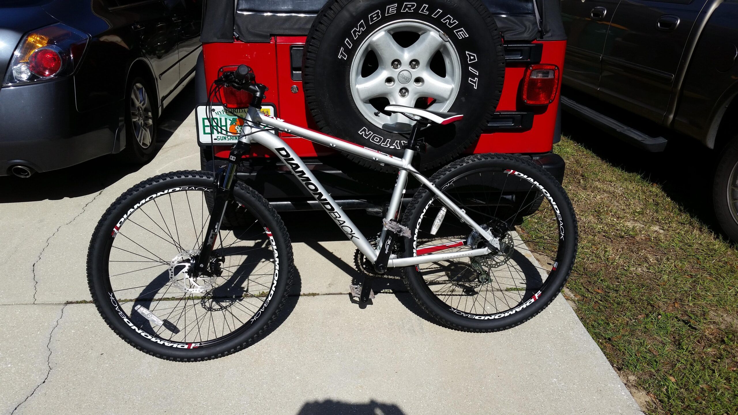 Diamondback Axis 27.5: A silver Diamondback mountain bike parked on a concrete driveway next to a red vehicle. The bike features thick tires and a simple frame design, with a view of the handlebars and seat. The background includes a gray car and some grass.