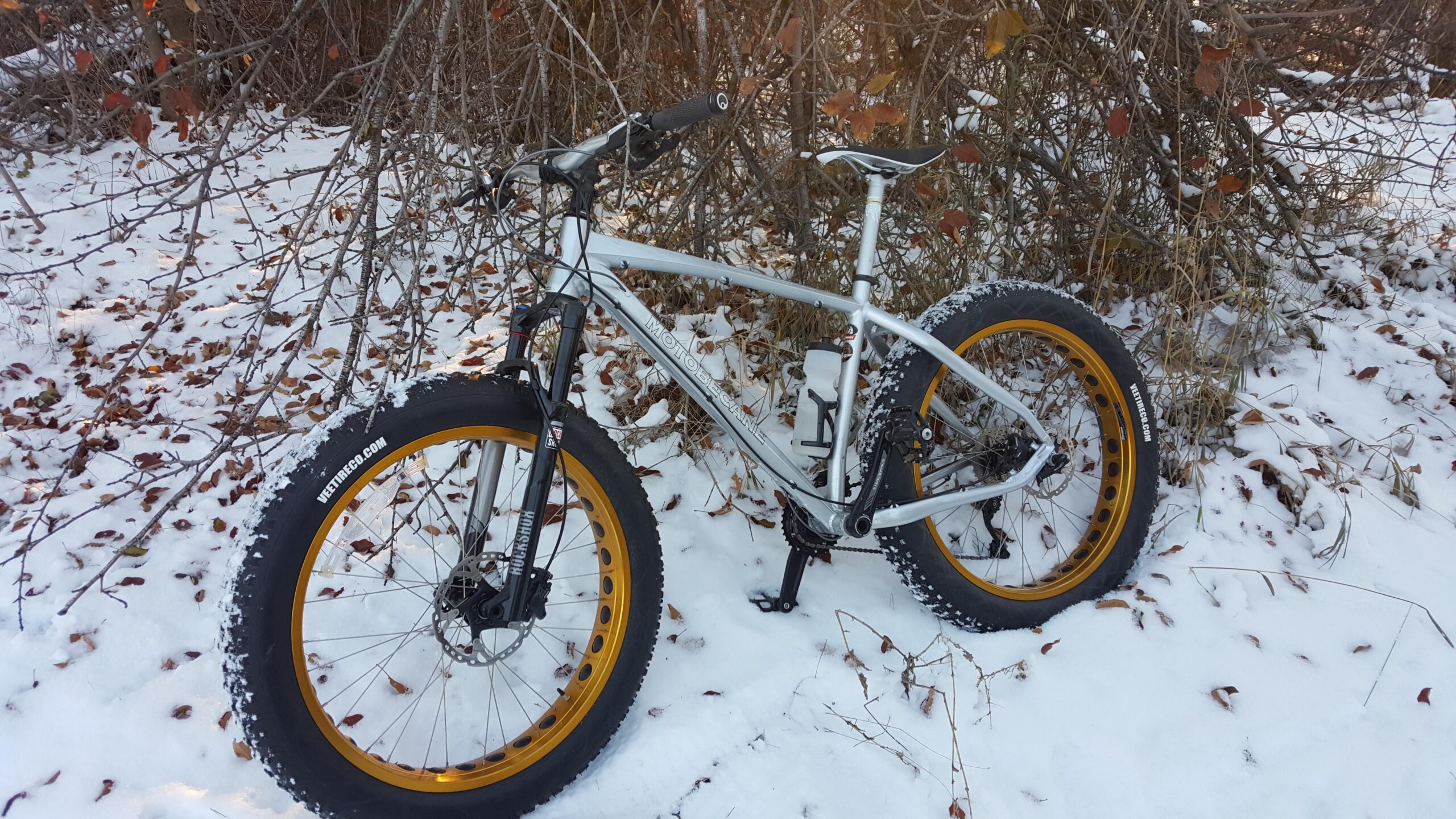 Motobecane Sturgis Bullet: A silver fat bike with orange rims is leaning against a bush, partially covered in snow and surrounded by fallen leaves. The scene depicts a wintry outdoor setting.