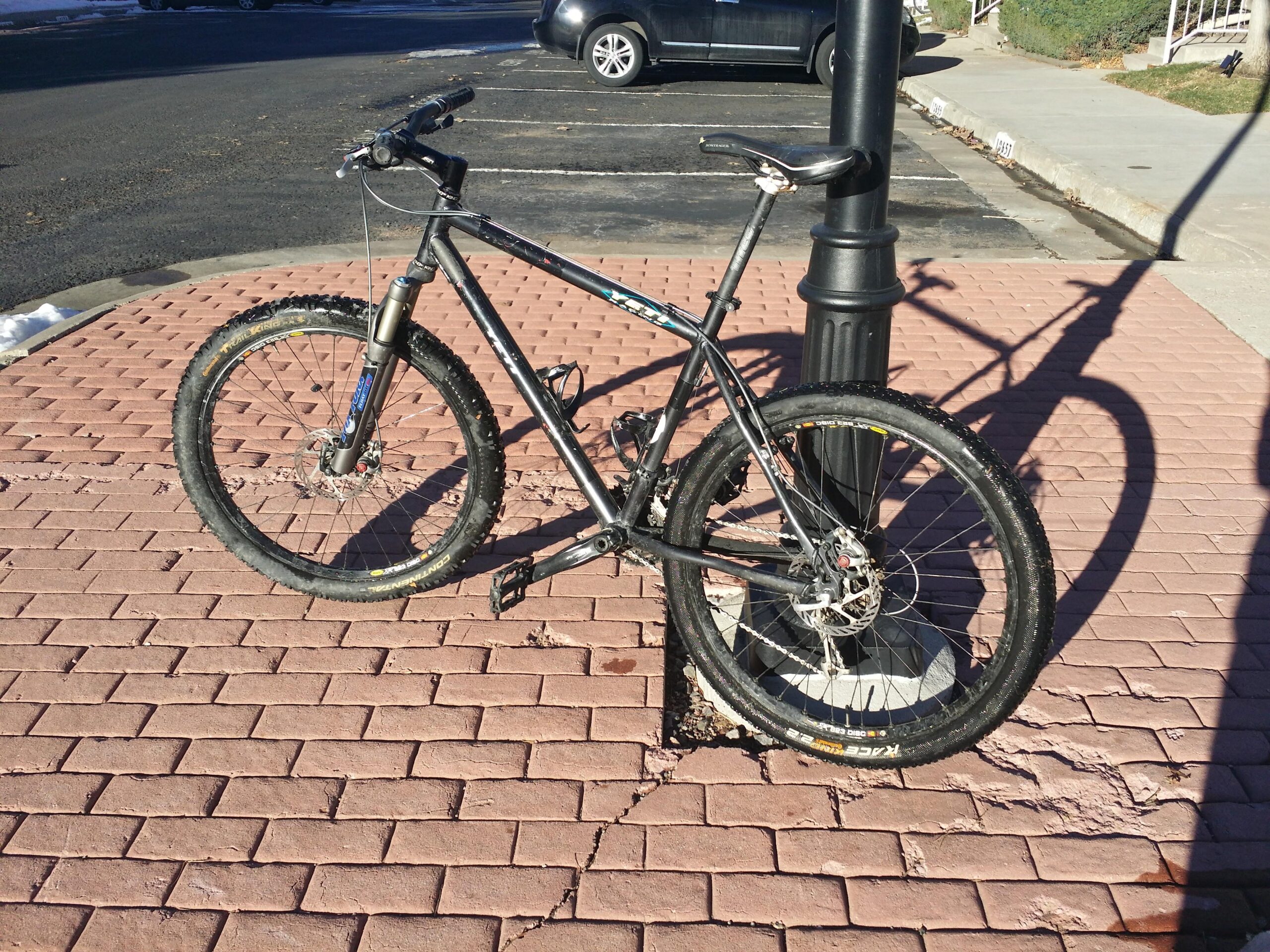 Kona Caldera: A black mountain bike leaning against a lamppost on a paver sidewalk, with its shadow casting on the ground. The bike features thick tires and a front suspension fork, set in a sunny outdoor environment.