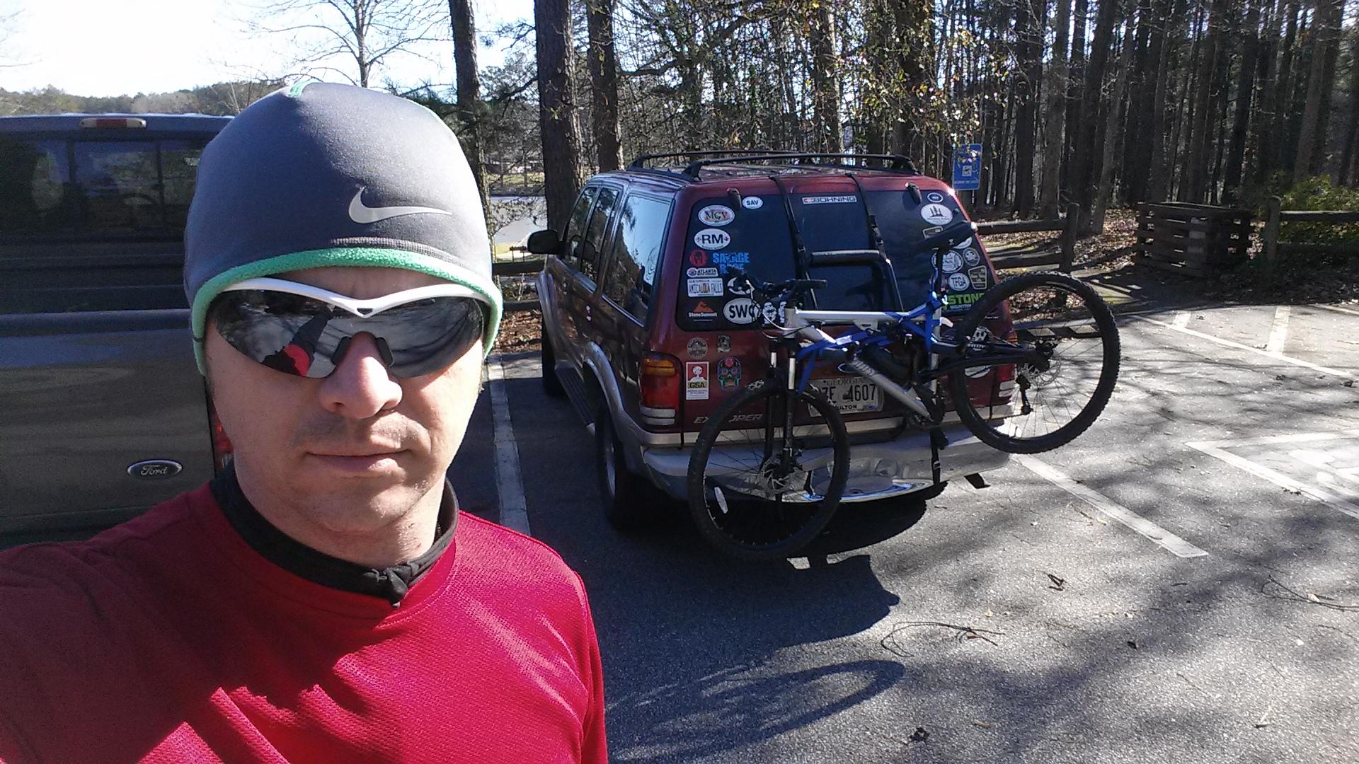 Gravity FSX 29One: A person wearing a gray beanie and sunglasses stands in front of a parked SUV with a mountain bike mounted on a bike rack. The background features trees and a clear sky, indicating an outdoor recreational location. The person is dressed in a red long-sleeve shirt, ready for a biking adventure.