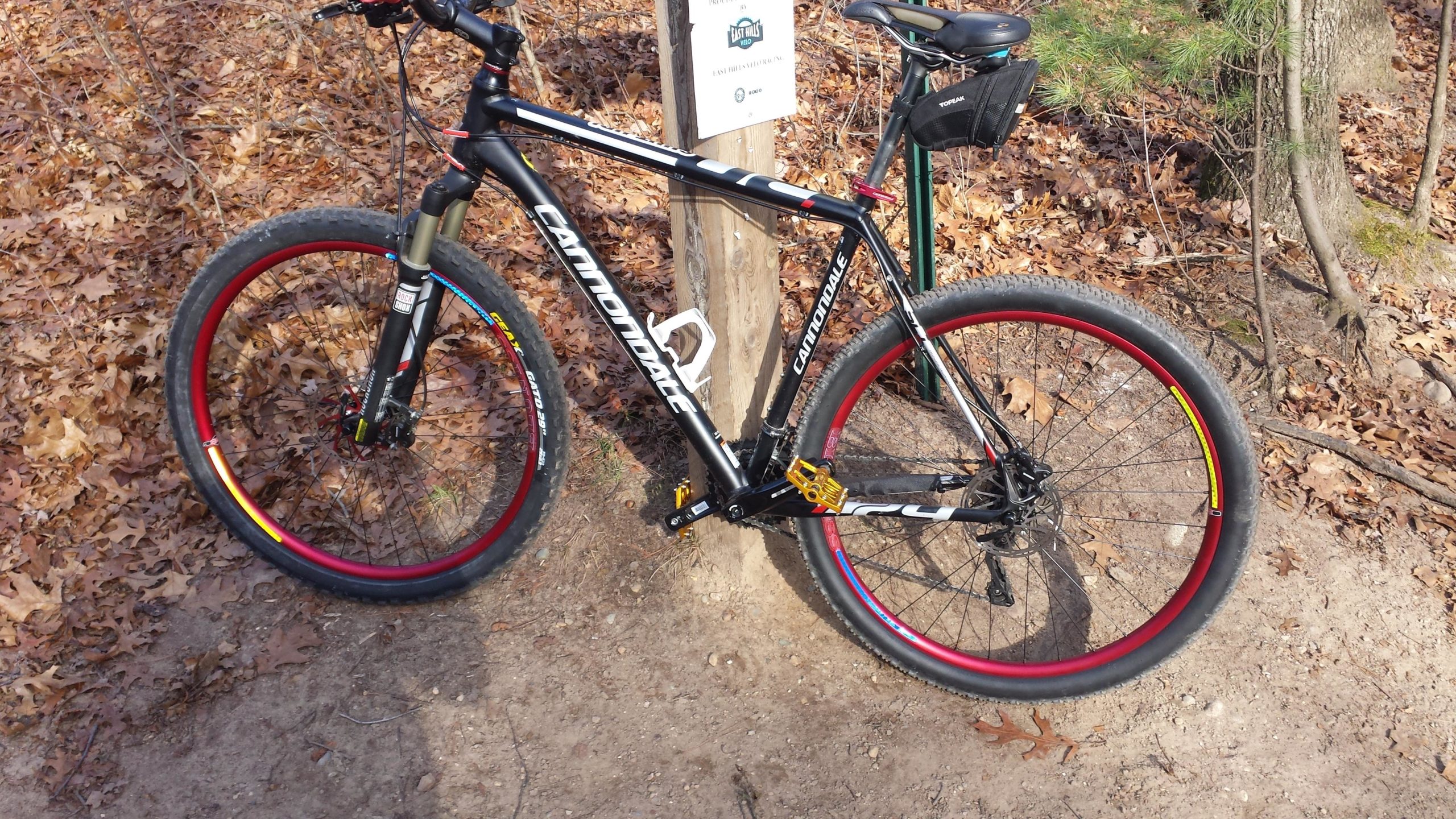 A black mountain bike with red and yellow accents parked next to a wooden trail sign, surrounded by fallen leaves and trees in a wooded area. Luton Park Trail mountain bike trail.