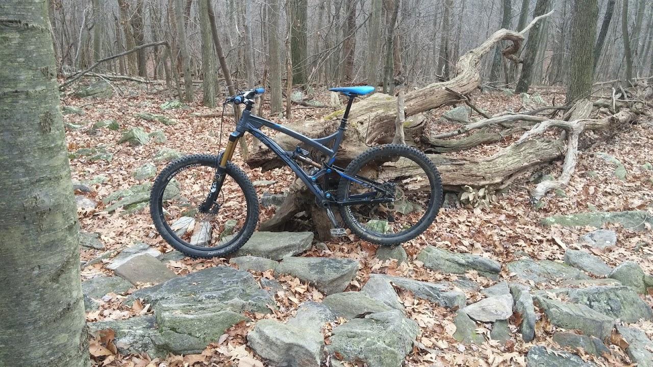 A mountain bike resting on rocky terrain surrounded by autumn leaves in a forest setting, with bare trees in the background and a fallen tree trunk nearby. Frederick Watershed mountain bike trail.