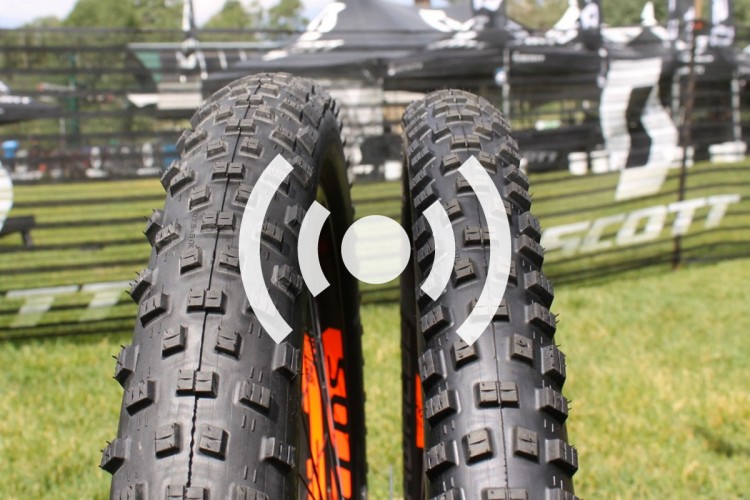 Two mountain bike tires positioned closely together, showcasing their detailed tread patterns. In the background, a blurred scene of a sporting event with branding and tents is visible.