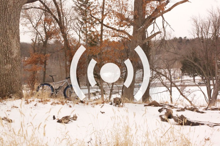 A winter landscape showing a bike leaning against a tree, surrounded by snow and dried grass. The scene captures a serene outdoor environment with a lake in the background and bare trees with some remaining autumn leaves. A graphic overlay of concentric circles is present, suggesting a theme of motion or connectivity.