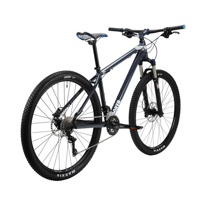 Charge Charge Cooker 29er Performance Exclusive: An image of a modern mountain bike featuring a dark blue frame, front suspension, and wide tires with a knobby tread suitable for off-road terrain. The bike is positioned at an angle that highlights its frame design and components.