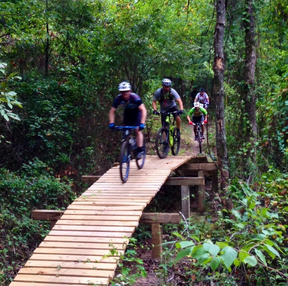 A group of four mountain bikers riding on a wooden bridge through a wooded trail, surrounded by lush greenery and trees. The riders are wearing helmets and cycling gear, showcasing the outdoor adventure and thrill of mountain biking. Bringle Lake Mountain Bike Trail System mountain bike trail.