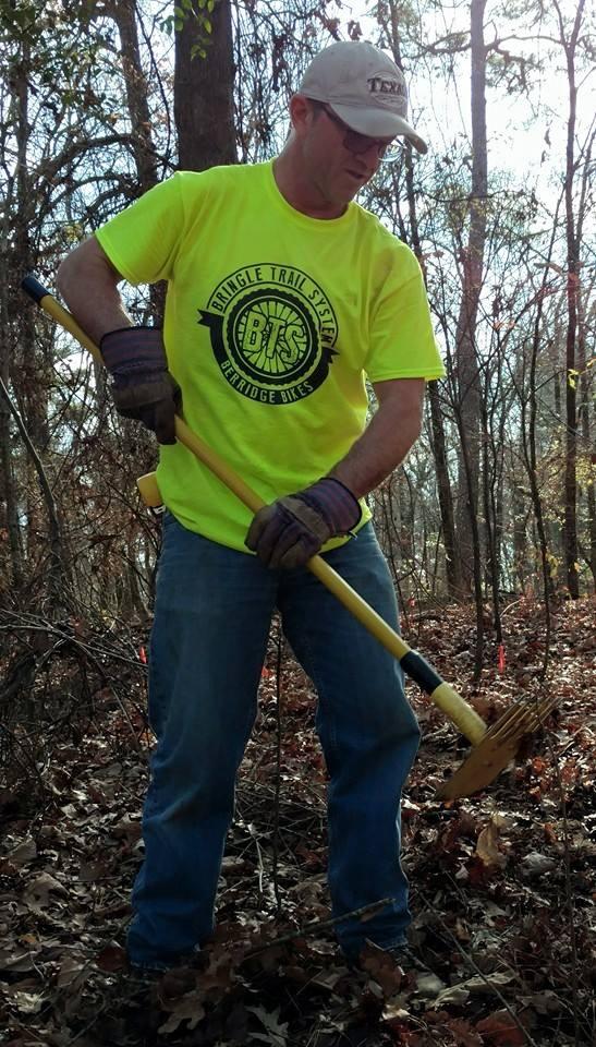 A person wearing a bright yellow shirt with a bicycle logo is using a gardening tool while standing in a wooded area covered with fallen leaves. They are wearing gloves and denim jeans, and are focused on their task among the trees. Bringle Lake Mountain Bike Trail System mountain bike trail.