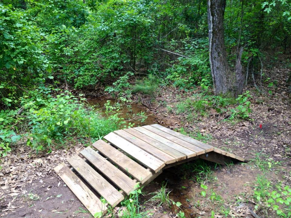 A small wooden bridge crosses over a shallow stream in a lush, green forest. Surrounding the bridge are various plants and trees, creating a tranquil, natural setting. The ground is covered with a mix of dirt and fallen leaves. Bringle Lake Mountain Bike Trail System mountain bike trail.