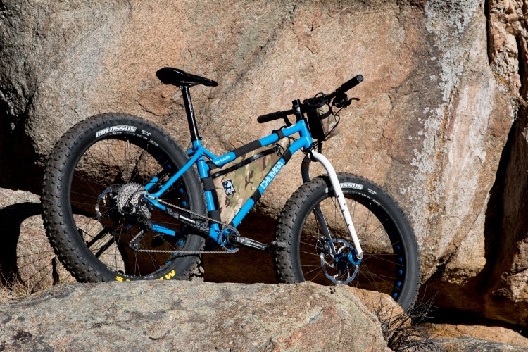 A blue fat bike with thick tires rests against a rocky surface. The bike features a camouflage-patterned frame bag and is positioned in an outdoor setting with natural stone formations in the background. Sunlight highlights the bike's details, showcasing its rugged design.
