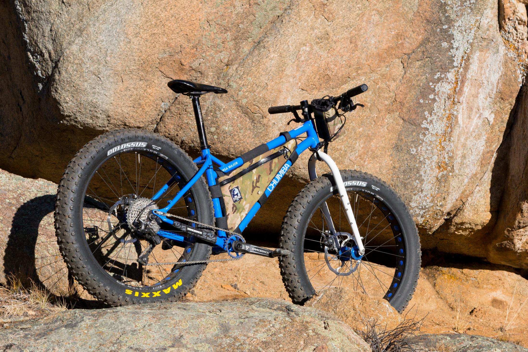 Chumba Ursa Major: A blue fat bike with large, rugged tires rests against a rocky backdrop. The bike features a camouflage frame bag and has a sleek design, suited for off-road adventures. Sunlight highlights the textures of the rocks and the bike's components.