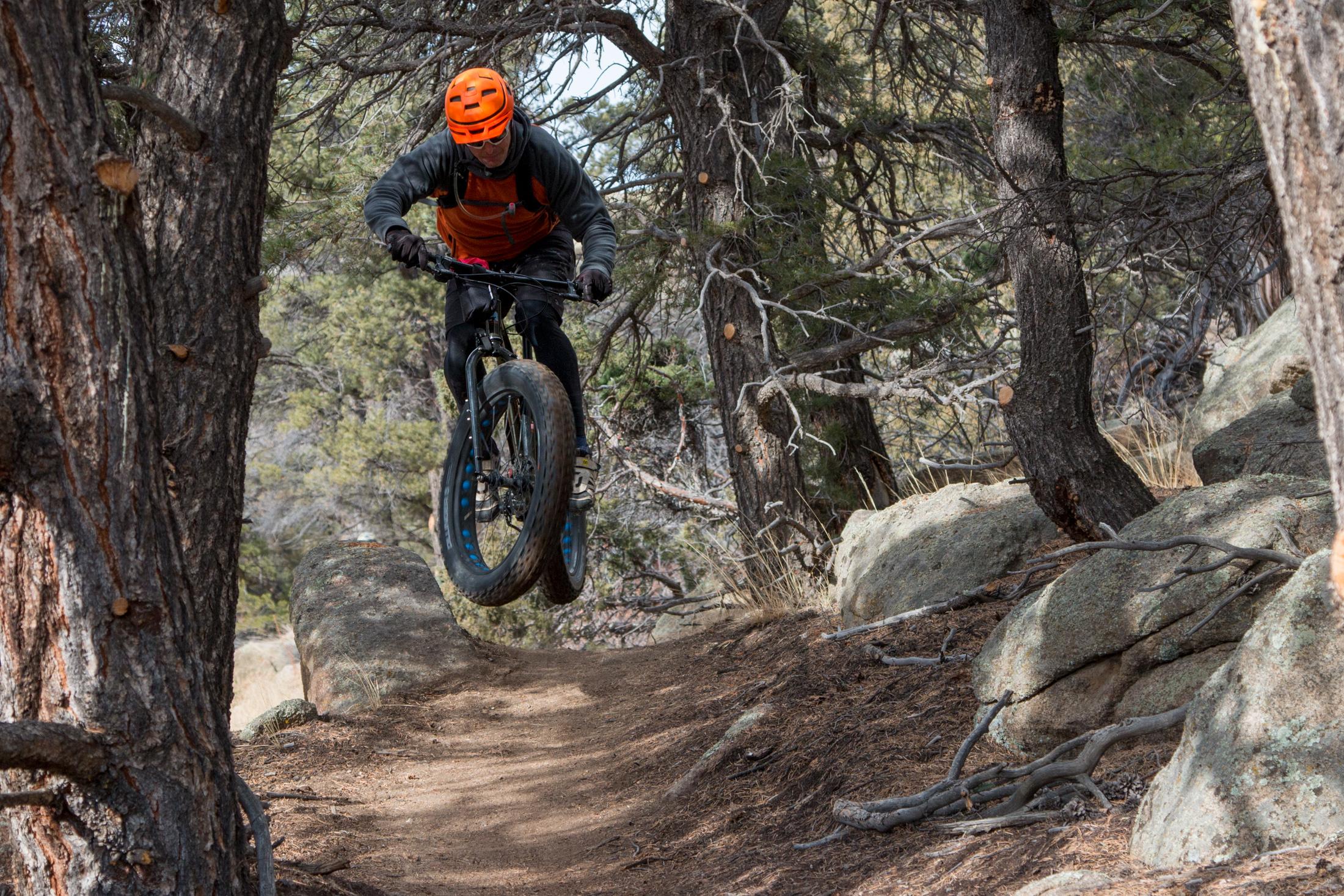 A mountain biker in an orange helmet performs a jump over a rocky trail surrounded by trees. The bike features wide tires, and the rider is dressed in a dark hoodie and black shorts. The scene captures the action and excitement of mountain biking in a natural setting. Midland Hills Trails mountain bike trail.
