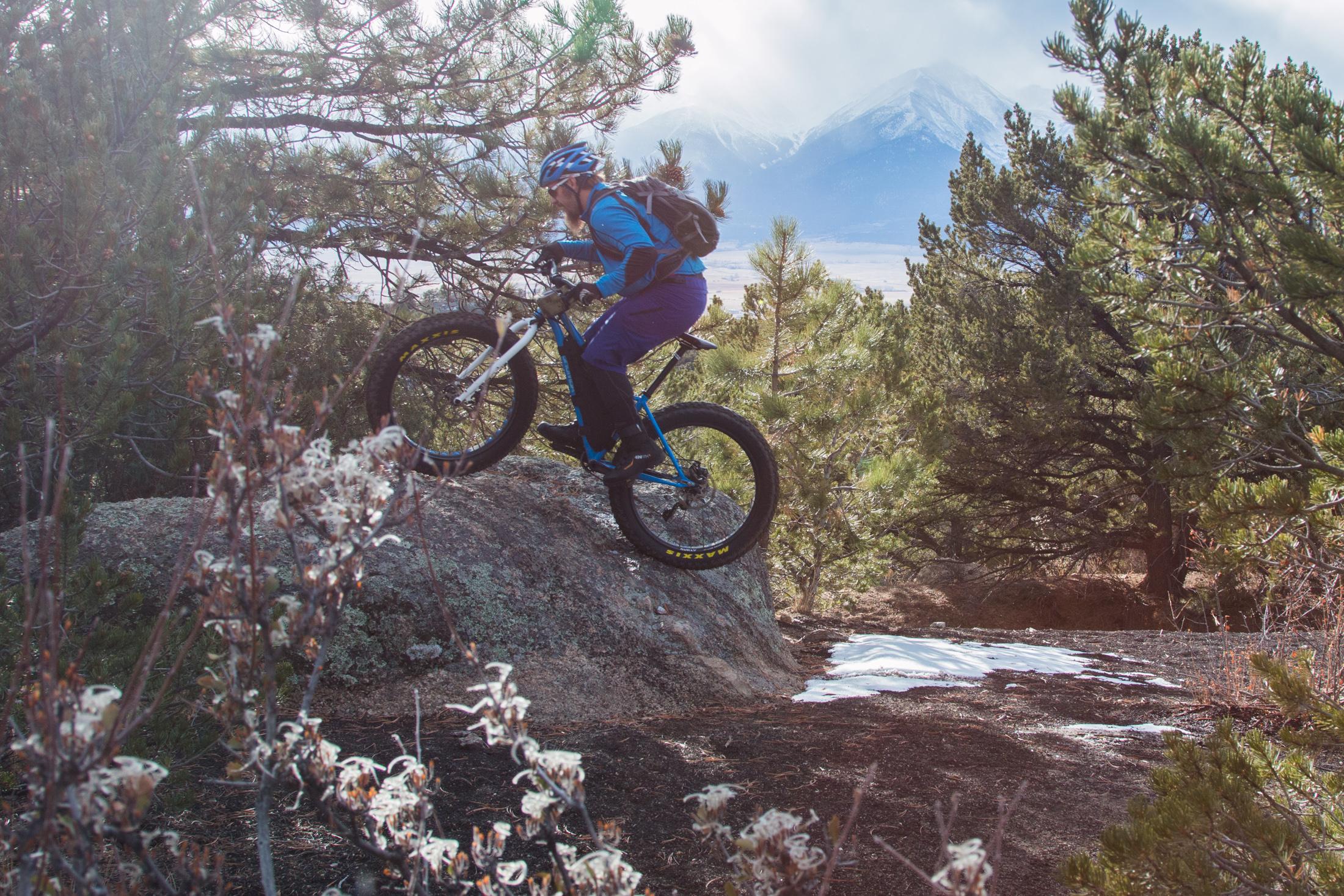 A mountain biker in blue gear ascends a rocky terrain surrounded by pine trees, with mountains and a hint of snow visible in the background. The rider is focused and balanced on the bike, showcasing a dynamic outdoor adventure scene. Midland Hills Trails mountain bike trail.