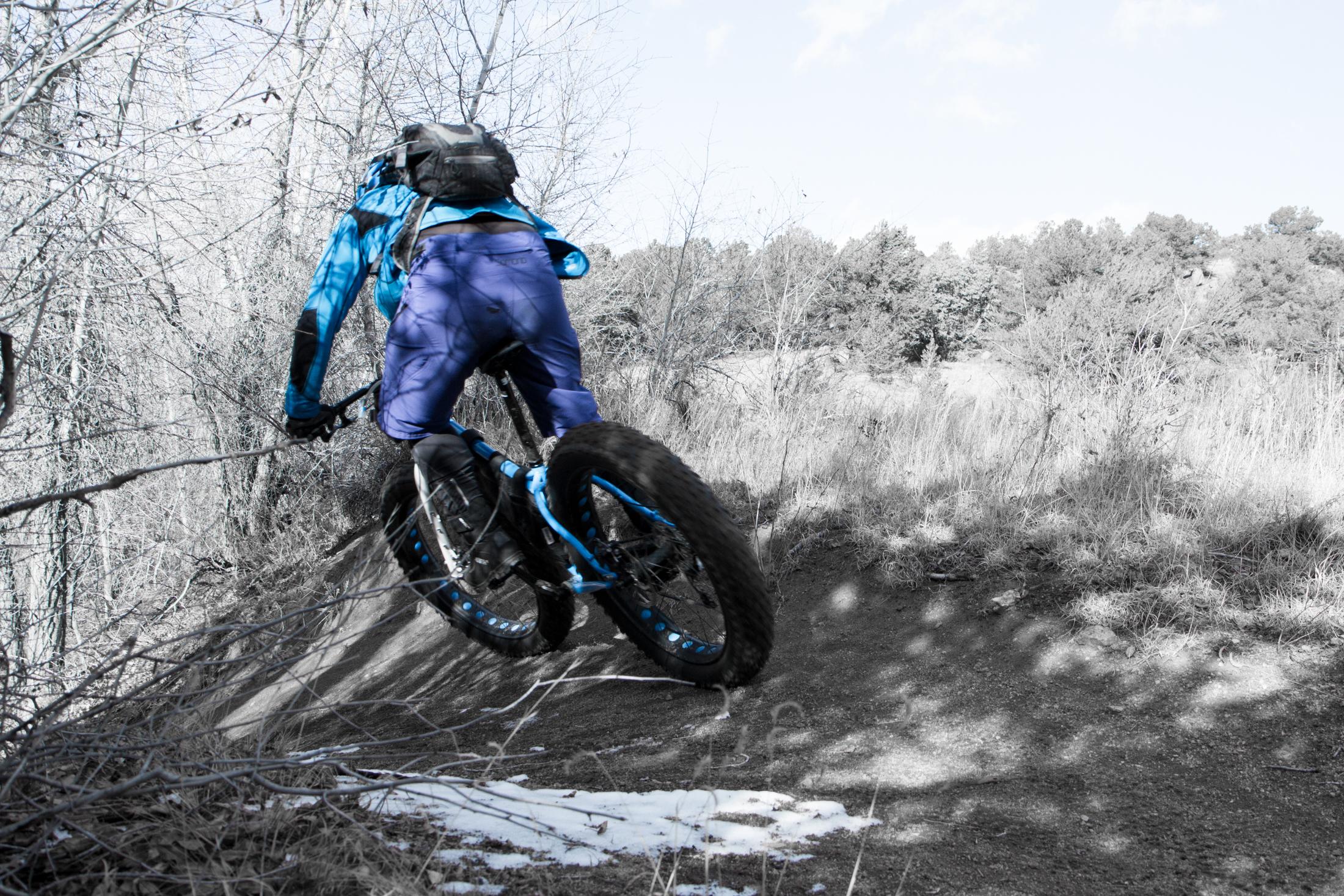 A mountain biker in a blue and black outfit is pedaling on a dirt path surrounded by trees and underbrush, with patches of snow visible on the ground. The cyclist is viewed from behind, emphasizing their movement and dynamic posture as they navigate the trail. Midland Hills Trails mountain bike trail.