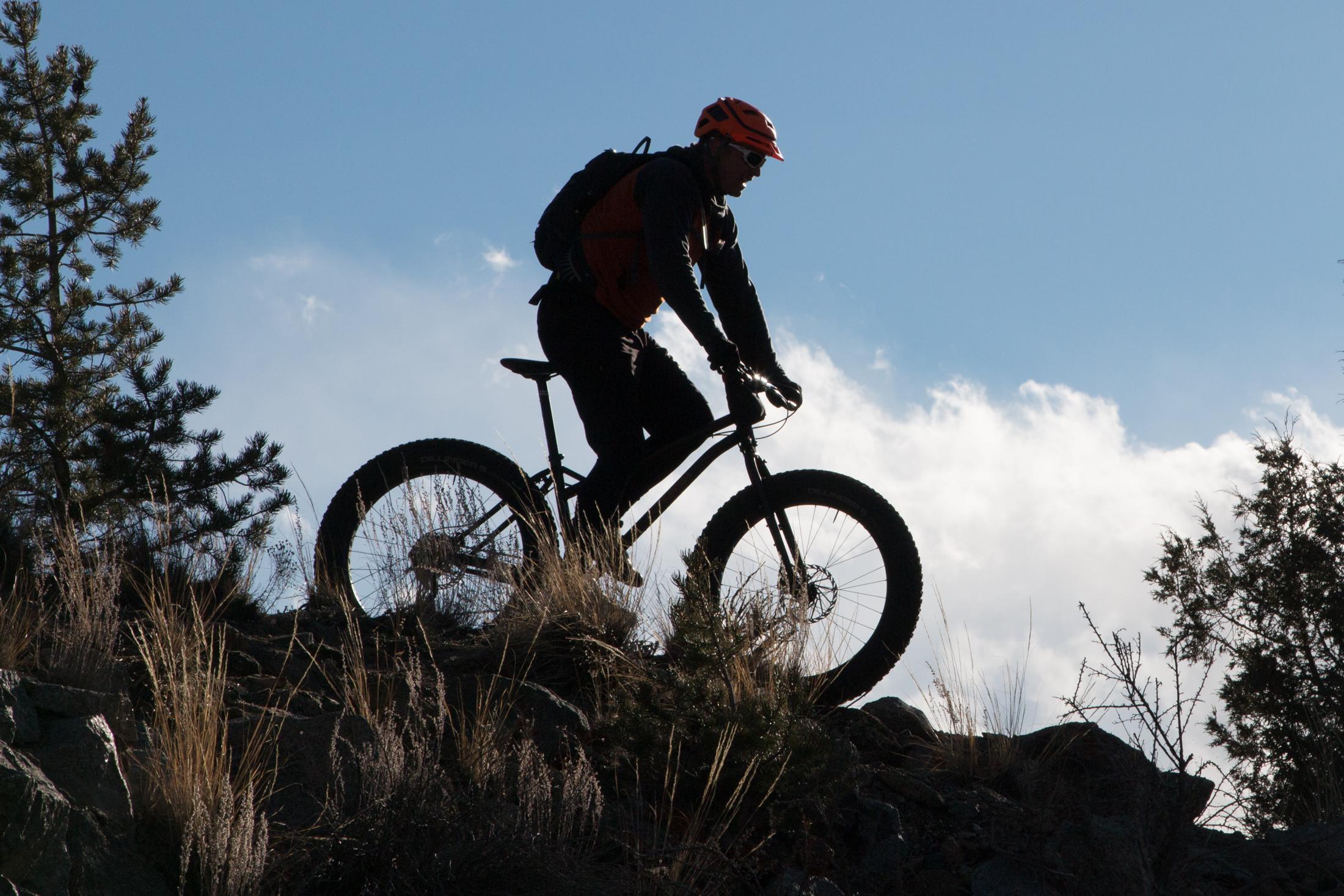 Silhouette of a mountain biker riding up a rocky trail against a blue sky with clouds. The cyclist is wearing an orange helmet and a backpack, surrounded by tall grass and pine trees. Midland Hills Trails mountain bike trail.