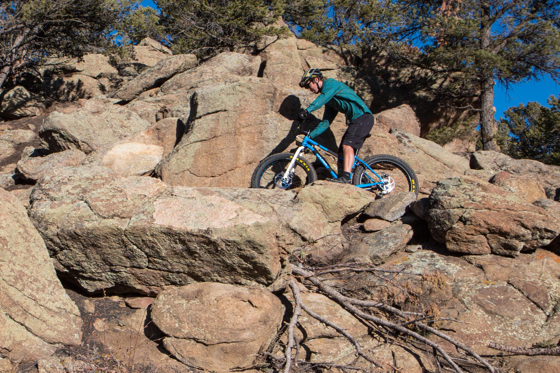 A mountain biker carefully navigates rocky terrain, balancing on large boulders under a clear blue sky. The cyclist wears a helmet and sports gear, showcasing an adventurous moment in outdoor biking. Pine trees are visible in the background. Midland Hills Trails mountain bike trail.