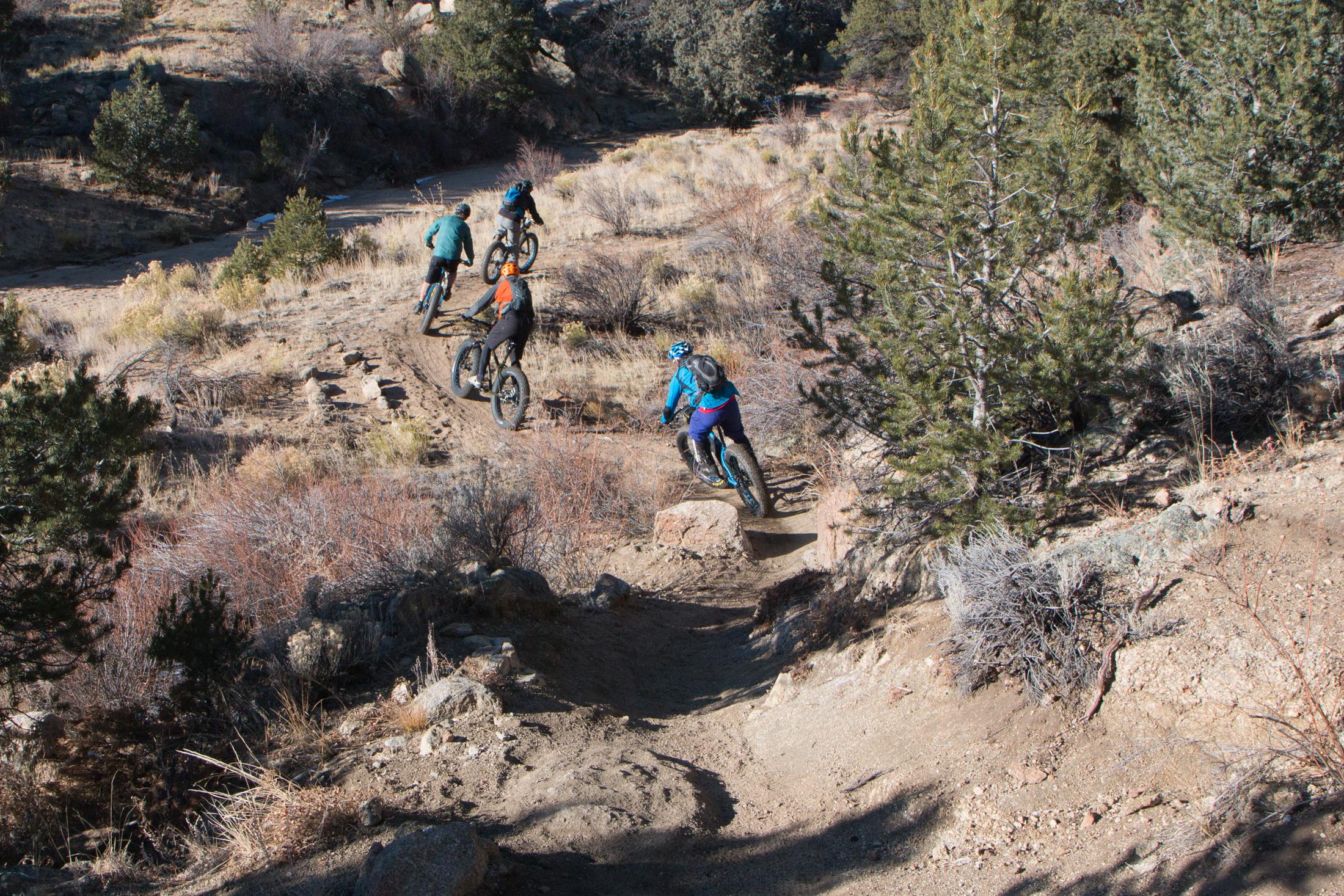 A group of four mountain bikers riding along a dirt path in a rugged outdoor landscape. The scene features rocky terrain, sparse vegetation, and trees, with the bikers wearing colorful gear as they navigate the winding trail. Midland Hills Trails mountain bike trail.