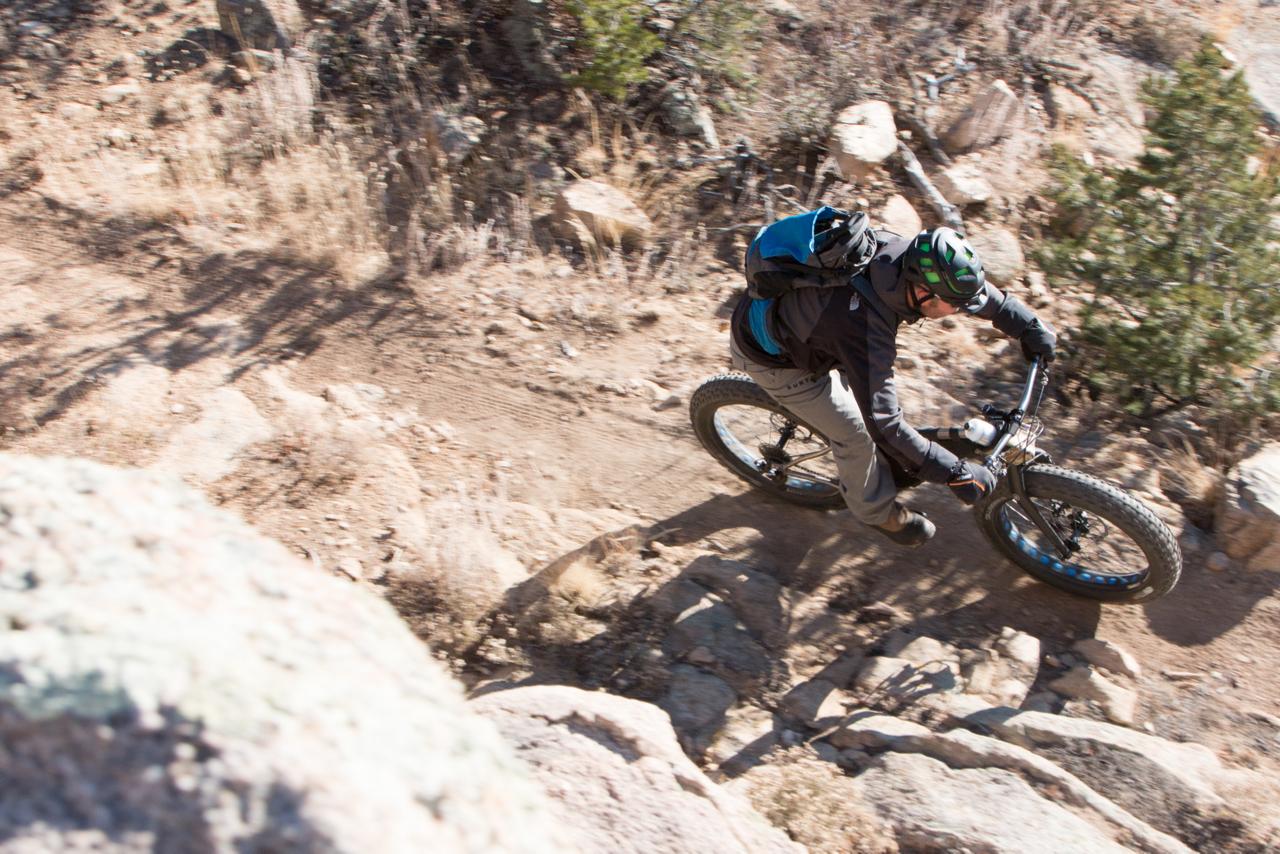 A person riding a mountain bike on a rocky trail, navigating through rough terrain with boulders and sparse vegetation in the background. The rider is wearing a helmet and a backpack, indicating a focus on outdoor sports and adventure. Midland Hills Trails mountain bike trail.
