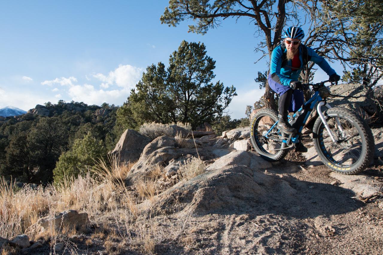 A mountain biker navigates a rocky trail surrounded by trees and rugged terrain under a clear blue sky. The cyclist wears a helmet and sunglasses, dressed in a bright blue jacket and pants, showcasing an adventurous outdoor scene. Midland Hills Trails mountain bike trail.
