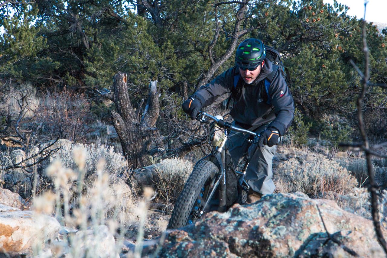 A mountain biker navigates rocky terrain in a dense forest, wearing a helmet and rugged outdoor clothing. The scene captures the cyclist leaning forward on a fat bike, with trees and vegetation in the background. Midland Hills Trails mountain bike trail.
