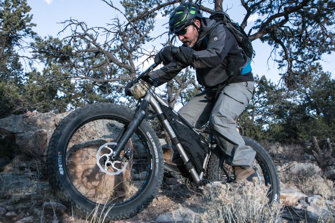 A cyclist riding a fat bike over rocky terrain, wearing outdoor gear including gloves, a helmet, and sunglasses. The background features sparse trees and shrubs against a clear sky. Midland Hills Trails mountain bike trail.