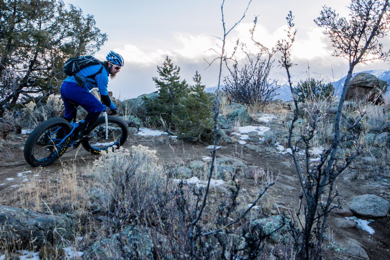A person riding a fat bike on a dirt trail surrounded by rocky terrain and sparse vegetation, with mountains and cloudy sky in the background. The rider is dressed in blue and wearing a helmet, focusing on navigating the trail. Midland Hills Trails mountain bike trail.