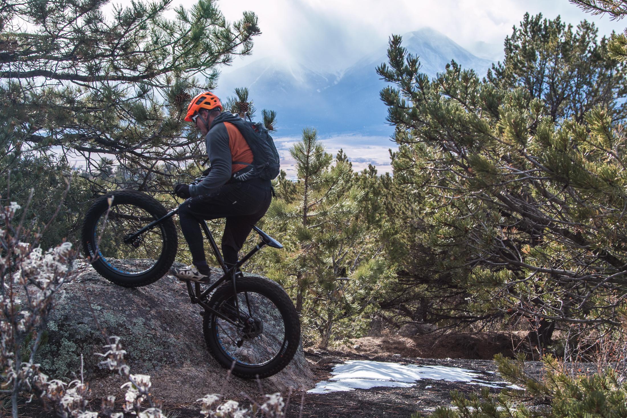 A mountain biker navigating a rocky trail surrounded by trees, with a partially snowy area in the background and mountains in the distance, under a cloudy sky. The cyclist is wearing an orange helmet and riding a fat bike, lifting the front wheel as they climb the rock. Midland Hills Trails mountain bike trail.