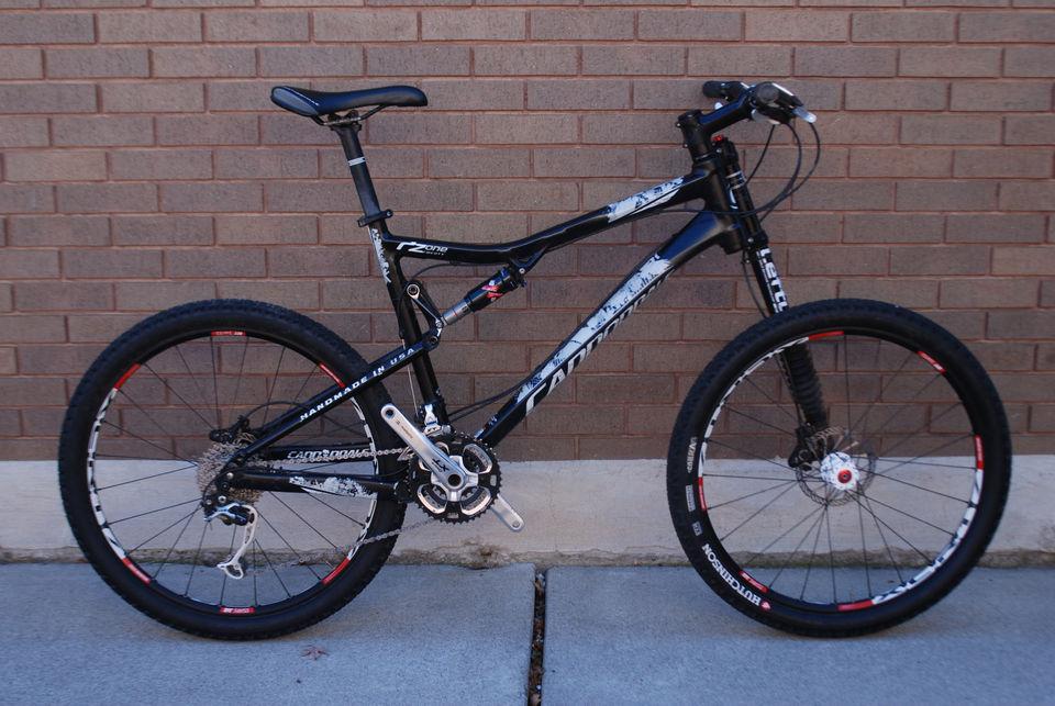 Cannondale RZ 120-1: A black mountain bike with a sleek frame, front suspension, and distinctive tire design, parked against a brick wall. The bike features red accents on the wheels and a prominent gear system. It is positioned on a concrete surface.