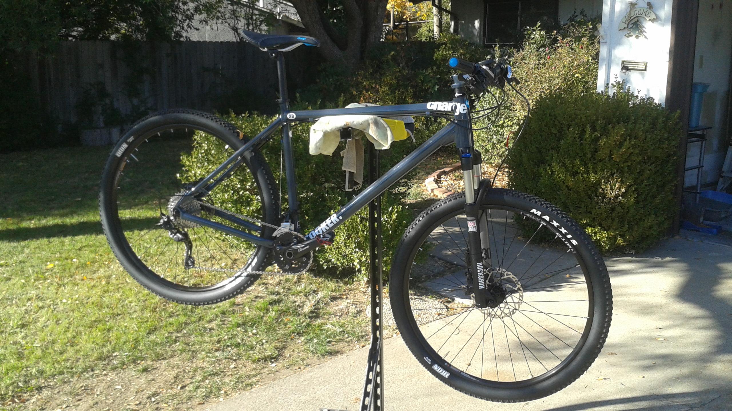 Charge Charge Cooker 29er Performance Exclusive: A mountain bike is suspended on a repair stand in a yard, with a towel draped over the frame. The bike features black tires and a blue and gray color scheme. In the background, there are green bushes and trees, and a welcoming home entrance is partially visible.