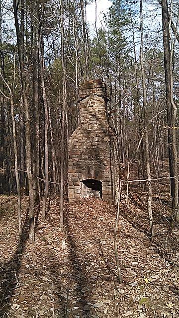 An old, weathered stone chimney standing alone amidst a forest of tall trees, with leaves scattered on the ground. The chimney shows signs of age and wear, and the surrounding area is peaceful, indicating that it's a remnant of a bygone structure. Sunlight filters through the trees, creating a serene atmosphere. Oak Mountain State Park mountain bike trail.