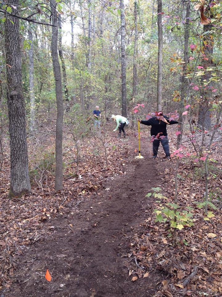 Three people are working on a dirt trail in a wooded area during autumn. One person is holding a measuring stick, while another is crouched down near the trail. The third person is standing further back. Colorful autumn leaves are sparse, and small orange flags are placed along the trail. The environment appears natural and lightly wooded, with trees in the background. Bringle Lake Mountain Bike Trail System mountain bike trail.