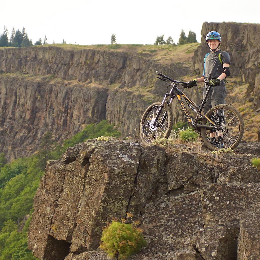 A young person wearing a blue helmet and protective gear stands proudly on a rocky cliff, next to a mountain bike. The landscape features rugged cliffs and green trees in the background, under a cloudy sky. Syncline mountain bike trail.